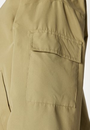 Khaki bunda s rukávy vybavenými čtvercovou náprsní kapsou, hladkou texturou a viditelným šitím. Materiál se zdá být lehký a odolný.