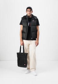 Melnā padded veste, melns T-krekls ar logo, bēšas bikses, baltas jaunās kedas, tur melnu auduma somu ar logo, attēlotas uz baltā fona.