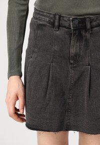 Gonna nera in denim con pieghe, orlo sfrangiato e cuciture in vista. Abbinata a un top aderente a maniche lunghe verde oliva.