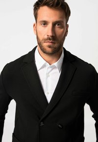 Blazer negro de doble botonadura en una tela suave con solapa de muesca, combinado con una camisa blanca de botones. Presenta un bolsillo en el pecho y un corte ajustado.