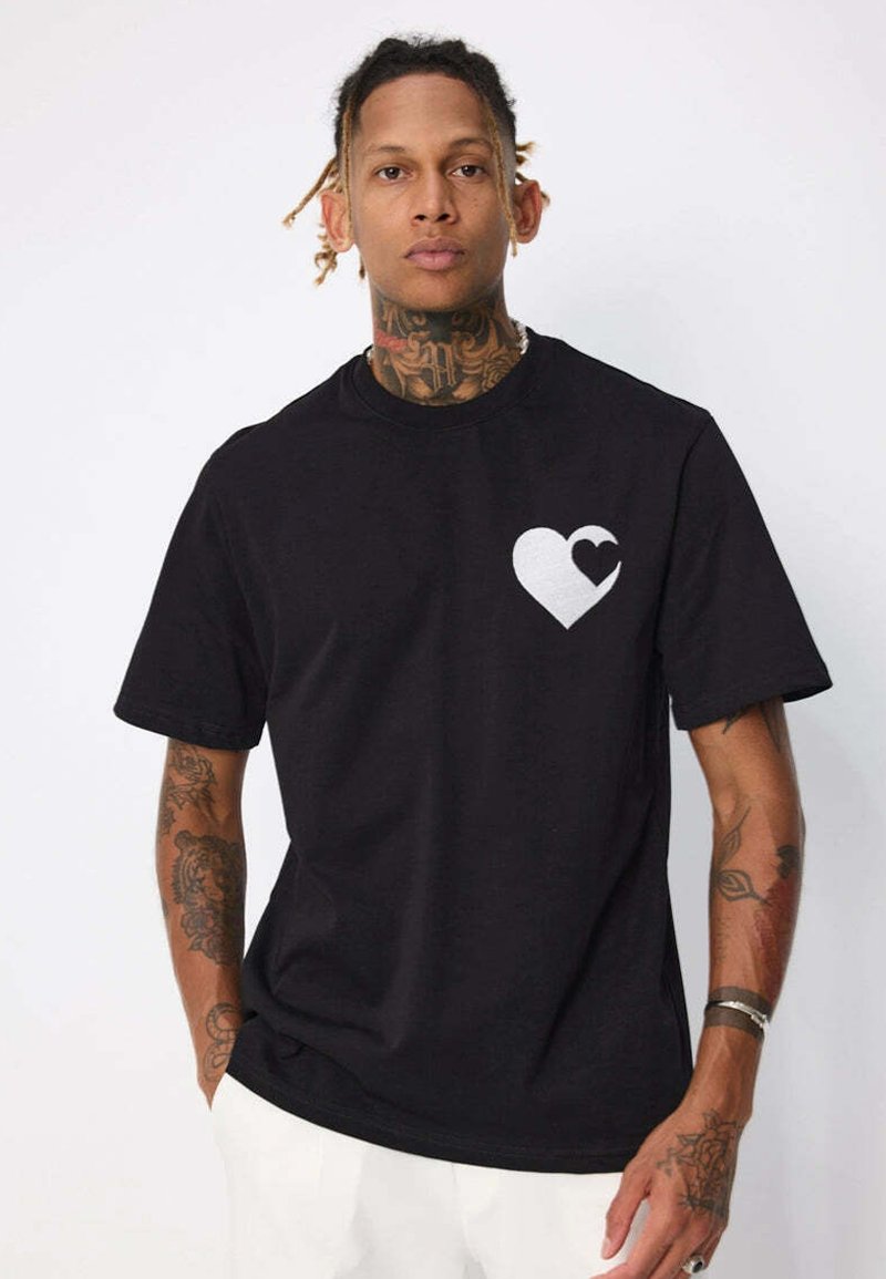 Homme avec des tatouages portant un t-shirt noir avec un cœur blanc et un plus petit cœur noir à l'intérieur, associé à un pantalon blanc, sur un fond uni.