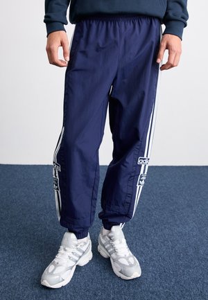 Osoba w granatowych spodniach dresowych Adidas z białymi paskami i białych butach sportowych Adidas stoi na niebieskim dywanie.