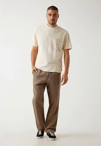 Maglietta grafica beige con design a logo circolare, abbinata a pantaloni marroni larghi e sneakers nere con suola bianca.