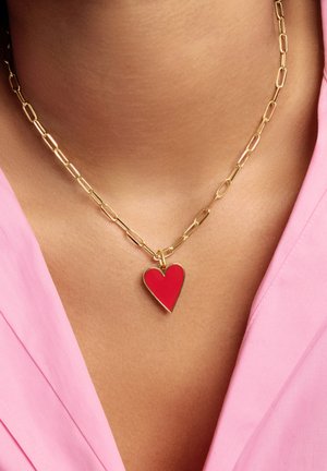 Collier en chaîne dorée avec de gros maillons tenant un pendentif en forme de cœur rouge, porté sur un haut rose à col en V.
