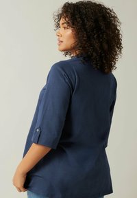 Top blu navy a tre quarti con una vestibilitàampia, caratterizzato da un dettaglio a bottone sulla manica e una texture liscia.