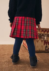 Röd plisserad kjol i plaid med gröna och gula accenter, kombinerad med blå texturerade strumpbyxor och svarta ankelboots med tofsdetaljer.