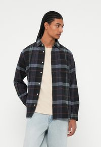 Sorte og marineblå ternet flannel skjorte med grønne og røde accenter, knaplukning, enkelt brystlomme og afslappet pasform over en beige t-shirt.