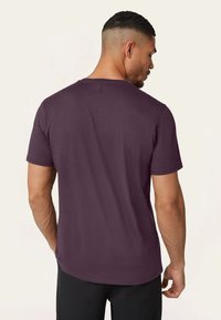 Man met kort haar in een nauwsluitend paars T-shirt en zwarte broek, staand met zijn rug naar de kijker gekeerd en zijn hoofd iets naar rechts gedraaid.