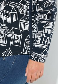 Marineblaues Langarmoberteil mit weißen Hausmotiven, runder Ausschnitt und abgerundetem Saum, kombiniert mit Jeans.