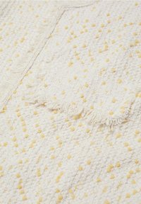 Tissu blanc texturé avec des points jaunes en relief, présentant un détail de frange le long d'un bord, créant une surface douce et tactile.