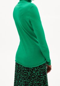 Långärmad, figurnära grön turtleneck av slätt tyg, ihop med en svart kjol med ett grönt blommönster.