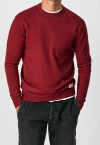 Röd stickad tröja med rund halsringning, ribbade ärmar och nederkant, mjuk textur, bärs över en vit skjorta, kombinerad med mörka byxor.