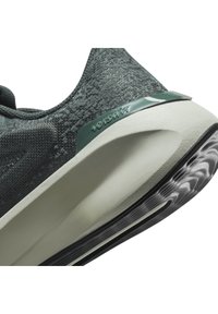 Zapato deportivo verde oscuro con parte superior texturizada, suela de goma gris y un detalle trasero liso con la inscripción "versait". Forma redondeada con líneas curvas.