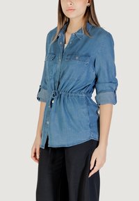 Camicia in denim azzurro chiaro con vita sagomata, chiusura frontale con bottoni, due tasche sul petto e maniche arrotolate, caratterizzata da una texture liscia e dettagli arricciati.