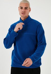Blauer Fleecepullover mit einem halben Reißverschlusskragen, langen Ärmeln und einer glatten, weichen Textur. Besitzt eine horizontale Naht über der Brust.