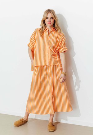 Femme blonde portant une chemise boutonnée orange et blanche à rayures verticales et une jupe midi assortie, debout contre un mur blanc avec des chaussures marron à enfiler.