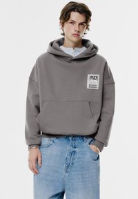 Grå oversized hoodie med en framficka och en märkespatch. Bärs med ljusblåa baggy jeans, som visar en avslappnad passform och mjuk textur.