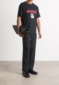 Mustat t-paita punaisella "HUGO" -tekstillä ja graafisella kuvalla, yhdistettynä mustiin housuihin ja kiillotettuihin mustiin kenkiin, pitäen kädessään rullattua mustaa laukkuasetusta.
