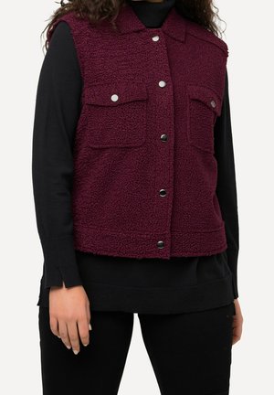 Femme portant un gilet sans manches en polaire bordeaux boutonné sur un haut noir à manches longues et un pantalon noir.