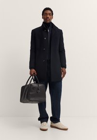 Czarny wełniany płaszcz z guzikami, w połączeniu z granatowymi dżinsami i białymi trampkami. Trzyma czarną skórzaną torbę z uchwytami. Prosty design.