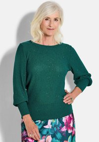 Grüner Strickpullover mit geripptem Saum und Ärmeln, der subtile Glitzerakzente aufweist. Das Modell kombiniert ihn mit einem Rock im Blumenmuster.