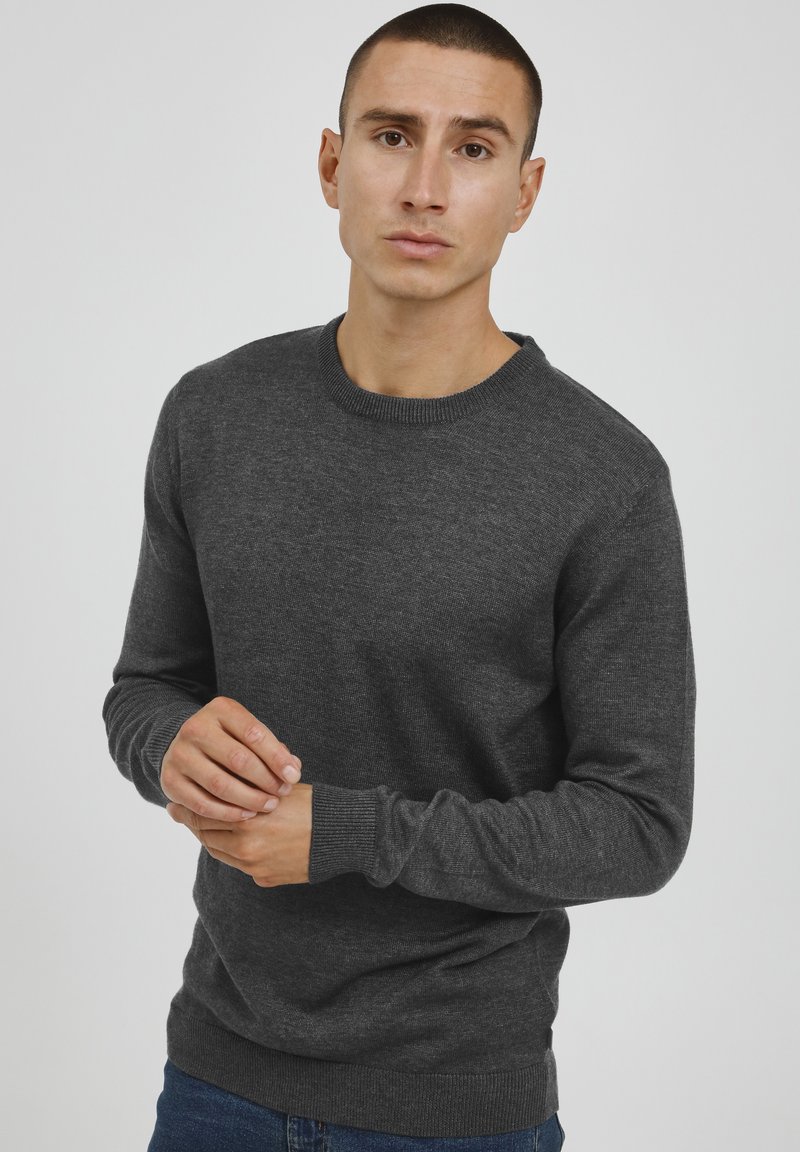 Donkergrijze gebreide trui met een ronde halslijn, geribbelde manchetten en een fitted taille, gedragen over blauwe jeans. Zachte textuur, minimaal ontwerp.