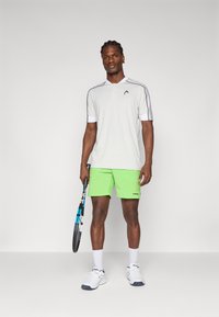 Polo gris con acentos blancos, pantalones cortos verdes y una raqueta de tenis. El atuendo presenta un corte ajustado y un diseño atlético.