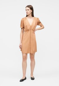 Μια γυναίκα στέκεται με το ένα χέρι στη μέση, φορώντας ένα κοντό, καφέ φόρεμα με φουσκωτά μανίκια, κουμπιά μπροστά και μαύρα επίπεδα παπούτσια, μπροστά σε ένα απλό φόντο.