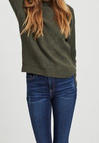 Pull en maille vert olive avec un col rond, associé à un jean skinny bleu foncé présentant un léger délavage et un design à cinq poches.