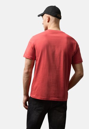 Mann, der ein schlichtes rotes T-Shirt mit kurzen Ärmeln, eine schwarze Kappe und schwarze Jeans trägt, von hinten vor einem einfarbigen hellen Hintergrund gesehen.