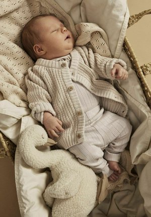 Beige gestrickte Strickjacke mit Holzknöpfen über einem hell gerippten Einteiler, begleitet von einem weißen plüschigen Schwan, der auf einer weichen cremefarbenen Decke ruht.