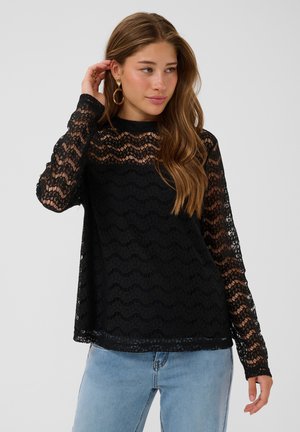 Femme aux longs cheveux bruns portant un haut noir en dentelle à manches longues et un jean bleu clair, se touchant les cheveux d'une main, regardant sur le côté.