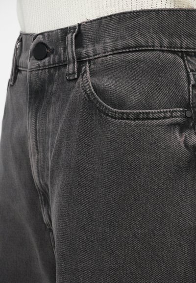 Nærbilde av mørkegrå denimjeans forfra som viser knapp, beltesløyfer og lommebrodering med en hvit strikket topp inni buksen.