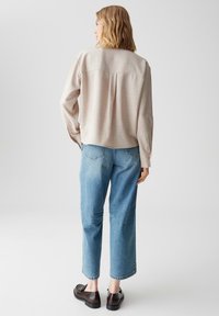 Camicia beige a maniche lunghe, texture; vestibilità rilassata con yoke posteriore. Abbinata a jeans a gamba dritta blu chiaro e mocassini scuri.