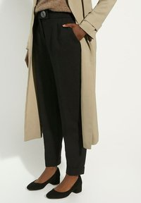 Pantalons noirs ajustés avec une taille haute et des poignets ourlés, associés à un long manteau beige et des chaussures à talons carrés en daim noir.