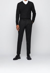 Černý svetr s výstřihem do V v kombinaci s upravenými černými kalhotami a černými botami, se hladkou texturou a strukturovaným designem.