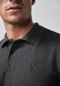 Polo gris oscuro con una tela texturizada, un cuello y tres botones negros. Presenta un logo en relieve en el área del pecho izquierdo.