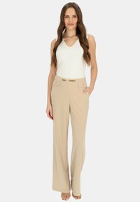 Beżowe spodnie o wysokim stanie z szerokimi nogawkami, z przednimi kieszeniami i połyskującym złotym łańcuchem na pasku. Połączone z białą bezrękawnik.
