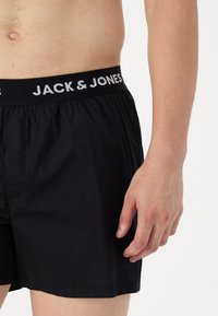 Črne bokserice z elastičnim pasom, na katerem je v belem napisu "JACK & JONES". Mehka tkanina, ohlapen kroj in brez vidnih vzorcev.