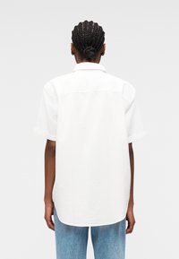 Personne aux cheveux tressés attachés en chignon, portant une chemise blanche à manches courtes et un jean bleu clair, vue de dos sur un fond uni.