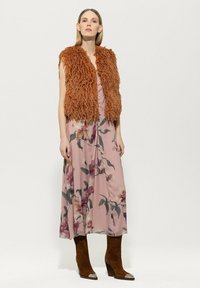 Braune, zottelige Fellweste über einem pinken Blumenkleid mit lila und grünen Akzenten; braune Stiefel mit Absatz und silbernen Zehenkappen.