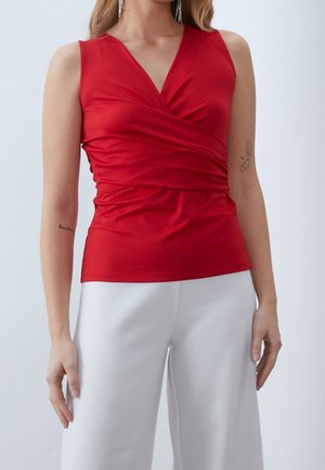 Kobieta ubrana w czerwoną, bezrękawnikową bluzkę z kopertowym dekoltem w kształcie litery V, zestawioną z białymi, szerokimi spodniami z wysokim stanem na jasnym tle.