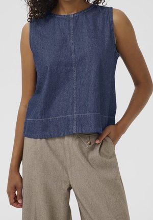 Femme portant un haut en jean bleu sans manches et un pantalon beige ajusté avec une poche boutonnée, main posée négligemment dans la poche.