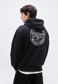 Νέος άνδρας με μαύρο φούτερ με κουκούλα, με λευκό λογότυπο αλόγων και κείμενο στην πλάτη, στέκεται πλάγια μπροστά σε ένα απλό φωτεινό φόντο.