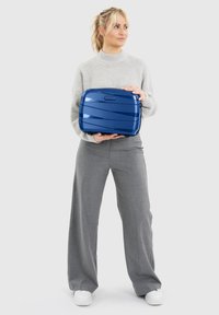 Vrouw met blond haar in een beige trui en grijze broek houdt een blauwe hard-shell koffer met het label "REDOLZ" tegen een grijze achtergrond.