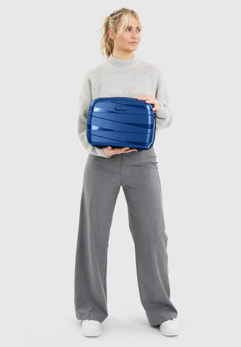 Vrouw met blond haar in een beige trui en grijze broek houdt een blauwe hard-shell koffer met het label "REDOLZ" tegen een grijze achtergrond.