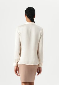 Cream blouse with a subtle textured pattern, long sleeves, and a rounded hem, paired with a fitted light brown skirt.