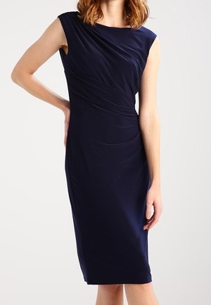 Femme portant une robe bleu foncé sans manches, longueur genou, avec des détails froncés sur le devant.