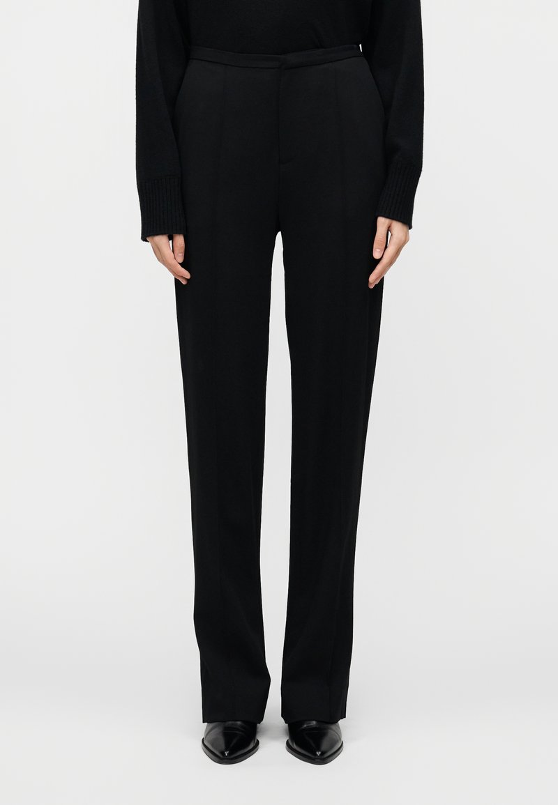 Pantalon noir, ajusté, à coupe droite, présentant une texture lisse et un pli subtil, associé à des chaussures noires à bouts pointus.