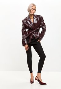 Blazer en cuir verni bordeaux avec larges revers, taille ceinturée, associé à des leggings noirs et des escarpins vernis bordeaux à bout pointu.